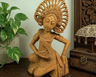 May include: A wooden sculpture of a Balinese dancer in a kneeling pose. The sculpture is carved from light-colored wood and features intricate details in the headdress, costume, and facial features. The dancer's expression is serene, and the overall effect is one of grace and elegance.