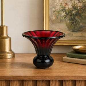 May include: A glass vase featuring a deep red, flared, fluted rim that transitions into a solid black, rounded base. The vessel has a smooth, reflective surface with vertical ridges extending from the centre toward the wide, circular opening.
