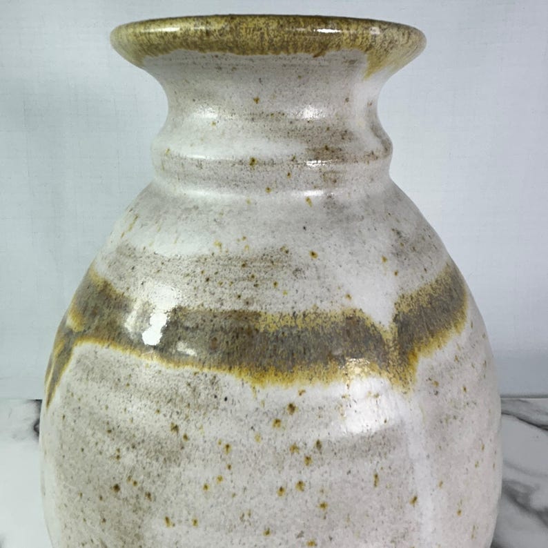 May include: A handcrafted ceramic vase with a wide body and a narrow neck. The vase features a speckled white glaze with bands of brown and yellow-brown around the body and rim. The vase has a glossy finish.