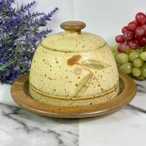 May include: A ceramic butter dish with a domed lid and a round base. The dish is beige with brown speckles and a floral design. The base and lid handle are brown. Grapes and lavender are in the background.