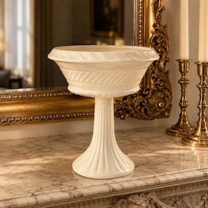 May include: A cream-coloured ceramic pedestal bowl featuring a wide, flared rim with a subtle diagonal ribbed texture. The bowl is supported by a slender, vertically fluted column base that widens at the bottom to provide a stable, circular foundation.