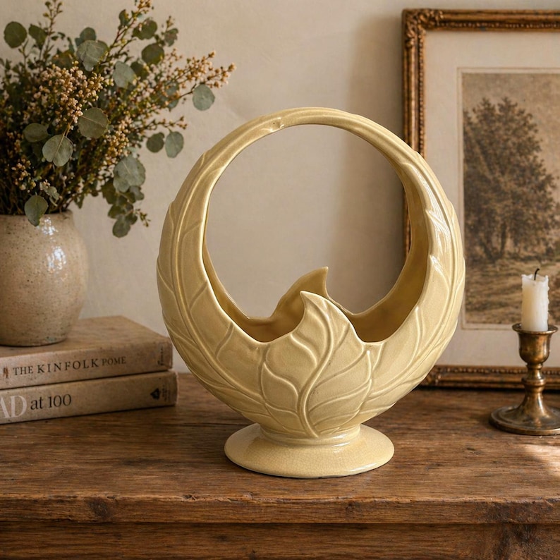 May include: A cream-colored ceramic basket with a large handle and leaf-like detailing. The basket sits on a round base and has a curved design. The basket is displayed on a wooden surface, with a vase of flowers and books in the background.