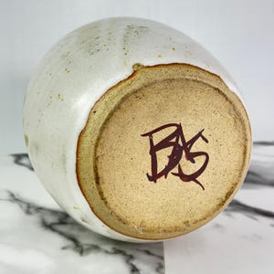 May include: A handmade ceramic vase with a white exterior and a brown rim. The base is a light brown color with dark brown lettering. The vase has a rounded shape and a textured surface.