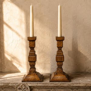 Peut inclure: Deux bougeoirs en bois brun foncé avec une base carrée et une colonne géométrique à plusieurs niveaux. Chaque bougeoir supporte une bougie effilée blanc cassé. Le grain du bois est visible, révélant un motif riche et texturé.