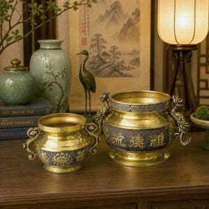 May include: Two gold-coloured incense burners with intricate dragon and floral designs. The larger burner has Chinese characters. The burners are displayed on a wooden surface, alongside other decorative items.