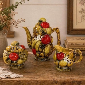 May include: A ceramic tea set featuring a teapot, creamer, and sugar bowl. The set is decorated with a fruit motif, including red apples, yellow pears, and green grapes. The pieces have a glossy finish and are arranged on a wooden surface.