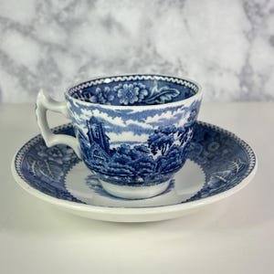 May include: Blue and white ceramic teacup and saucer set. The cup and saucer feature a detailed landscape scene with a church and trees. The rim of the cup and saucer have a decorative pattern.