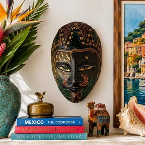 May include: An oval-shaped wooden wall mask featuring a dark base with intricate gold, green, and red painted patterns. The mask displays stylised facial features including almond-shaped eyes, a defined nose, and a small mouth with floral motifs.