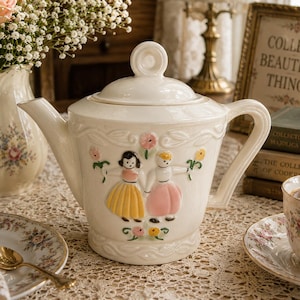 May include: Cream-coloured ceramic teapot with a lid, handle, and spout. The teapot features a decorative relief design with two figures in dresses and floral accents. A matching teacup and saucer are also visible.