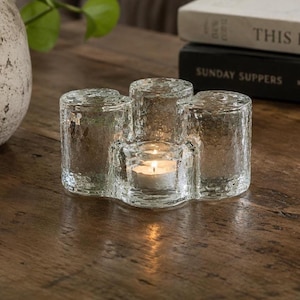 May include: A clear glass candle holder with four cylindrical compartments, one holding a lit tea light. The holder has a textured surface and sits on a wooden table. Books are in the background.