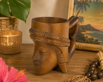 Puede incluir: Una taza de madera marrón tallada con un diseño de rostro y un asa en forma de mano. La taza está sobre una superficie de madera, con una imagen de temática hawaiana en el fondo. Una flor de hibisco rosa está en primer plano.