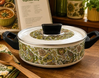 May include: A round white enameled metal pot with a lid, featuring a detailed green, brown, and yellow paisley floral pattern. The pot has two black side handles and a black knob on the lid. A wooden spoon rests on a matching patterned cloth nearby.