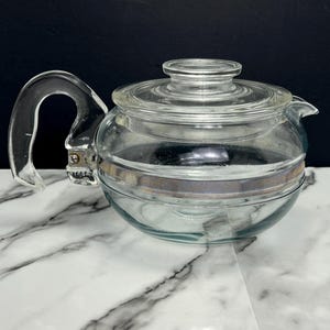 May include: A clear glass teapot with a metal band around the middle. The teapot has a lid and a handle.