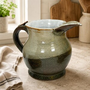 May include: A ceramic pitcher featuring a two-tone glaze with a light cream-coloured upper section and a dark, speckled forest green lower section. The vessel has a wide, rounded body, a curved handle, and a prominent pouring spout with a smooth finish.