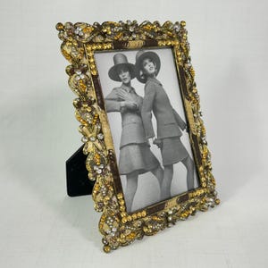 May include: An ornate gold picture frame with a black velvet easel back. The frame is decorated with amber and clear crystals and a brown border. The black and white photo shows two women in hats and suits.
