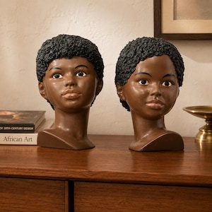 May include: Two identical busts of a person with dark brown skin and short, textured black hair. Each sculpture features a smooth, rounded face with dark eyes, defined eyebrows, and light-colored lips, capturing the head and neck in a portrait style.