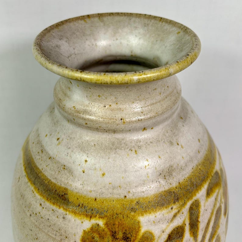 May include: A ceramic vase with a wide, slightly flared rim. The vase has a speckled, off-white base color with a band of yellow-brown around the middle and floral designs. The rim is a lighter shade of off-white.