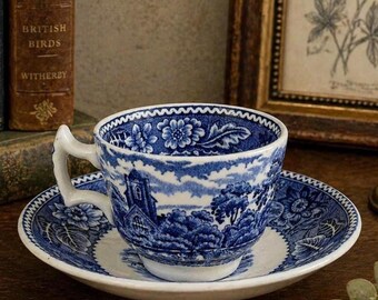 May include: A blue and white teacup and saucer set. The cup and saucer feature a detailed landscape design with floral and architectural elements. The handle of the cup is white. The set is placed on a wooden surface.