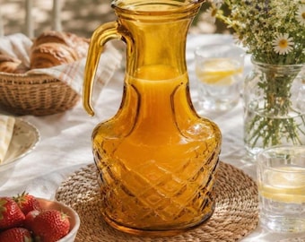May include: A vintage-style amber glass pitcher filled with orange liquid, likely juice, sits on a woven placemat. The pitcher has a diamond pattern and a curved handle. Strawberries and flowers are also on the table.