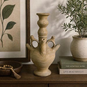 May include: A beige ceramic vase with three handles and a bulbous base. The vase has a unique, layered design with a central spout. The background includes a framed botanical print, a wooden bowl with beads, and a small potted plant.