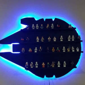 May include: A black wall shelf shaped like the Millennium Falcon from Star Wars, with blue LED lights around the edge. The shelf is filled with small Star Wars Lego minifigures.