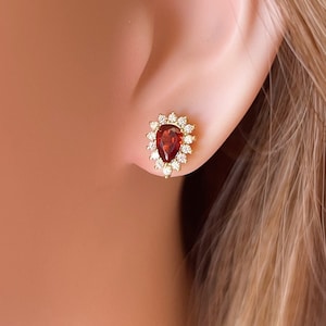 May include: A pair of gold earrings featuring a teardrop-shaped red gemstone surrounded by a halo of smaller white gemstones.