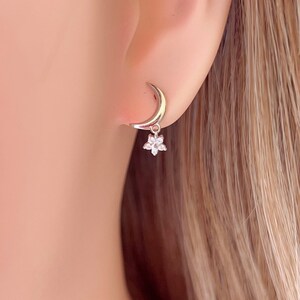 May include: Silver crescent moon hoop earrings with a small flower charm dangling from the bottom. The flower charm is set with clear cubic zirconia stones.