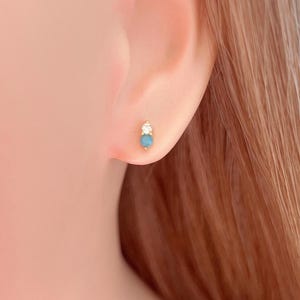 Puede incluir: Un pendiente de oro con una pequeña piedra redonda de color turquesa en la parte inferior y una piedra clara y brillante encima. El pendiente se muestra en una oreja.