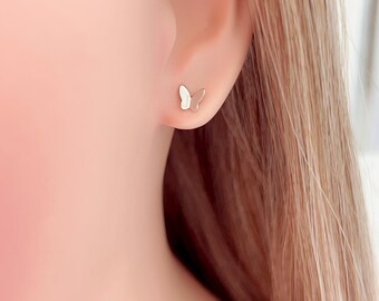 May include: A close-up of a person's ear with a small butterfly stud earring. The earring is silver-colored with a white wing design. The background is blurred, focusing on the earring.