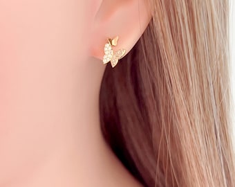 May include: A pair of gold-colored butterfly stud earrings. The earrings feature a detailed butterfly design, with the wings and body adorned with small, sparkling crystals. The earrings are shown on a person's ear.