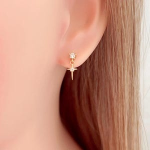 May include: A gold-colored earring with a starburst design. The earring features a small, clear gemstone at the top, a star-shaped pendant, and a dangling, pointed element. The earring is worn on a person's ear.
