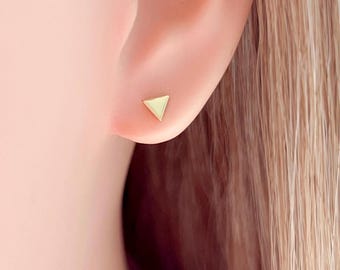 May include: A close-up of a person's ear with a small, gold-colored triangle stud earring. The earring is a simple geometric design, and the ear is in focus against a blurred background of skin and hair.