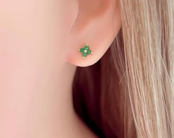Puede incluir: Primer plano de un pendiente de oro con un diseño de trébol de cuatro hojas. Cada hoja está hecha de pequeñas piedras redondas de color verde esmeralda. El pendiente está colocado contra la oreja de una persona y cabello rubio.