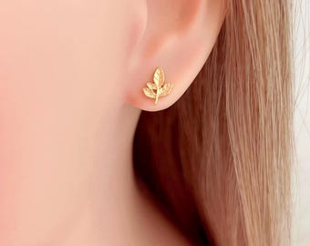 Könnte beinhalten: Nahaufnahme eines goldfarbenen Ohrsteckers in Blattform an einem Ohr. Der Ohrstecker ist eine detaillierte Miniatur eines Blattes, das seine Adern und Form zeigt. Der Hintergrund ist unscharf und hebt das Design des Ohrrings hervor.
