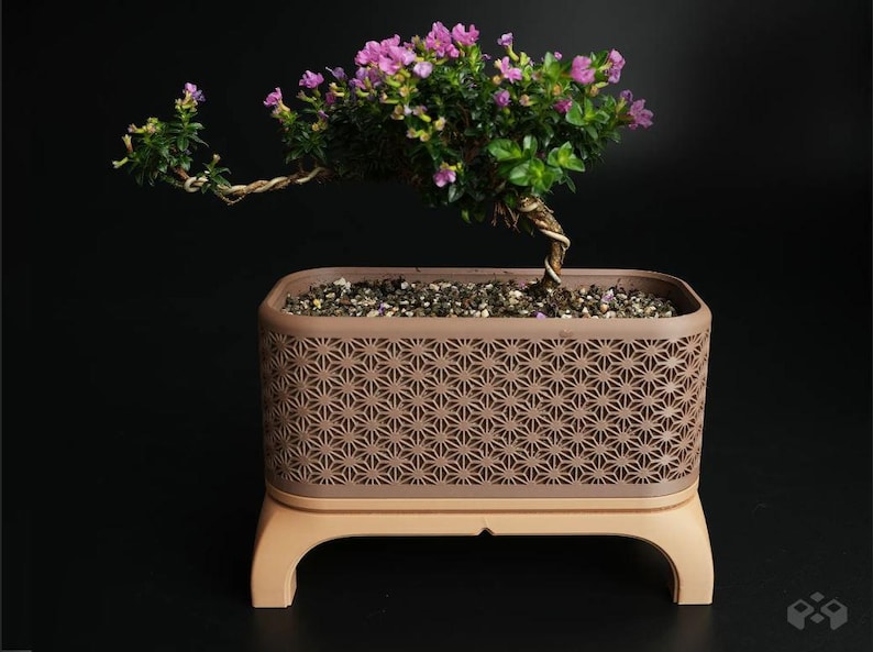 May include: A bonsai tree with pink flowers and green leaves in a decorative brown planter. The planter has a geometric pattern and sits on a light brown base. The tree's trunk is twisted, and the background is black.