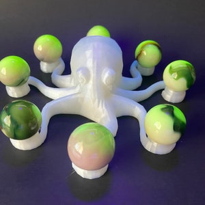 Può includere: Una scultura di polpo bianco con otto biglie verdi e bianche disposte attorno. Le biglie hanno un motivo a spirale e sono tenute da piccoli supporti bianchi. Il polpo è stampato in 3D e poggia su una superficie blu scuro.