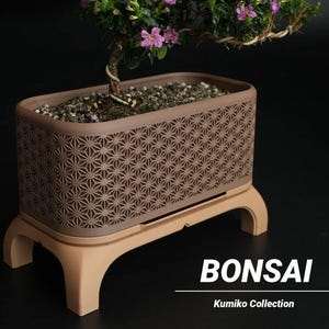 Może przedstawiać: Brązowa prostokątna donica do bonsai z geometrycznym wzorem, spoczywająca na jasnobrązowej podstawie. Donica zawiera ziemię i małe drzewko bonsai z różowymi kwiatami. Słowo "BONSAI" jest wyświetlane białym tekstem.
