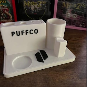 May include: A white plastic organiser with the word "PUFFCO" printed on the front. The organiser has compartments for storing various items, including a round container, a tall cylinder, and a small rectangular compartment. The organiser is sitting on a wooden surface.