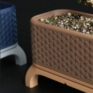 May include: A brown rectangular planter with a geometric starburst pattern, filled with soil and small stones. The planter sits on a matching base with curved legs. A blue planter with a similar design is visible in the background.