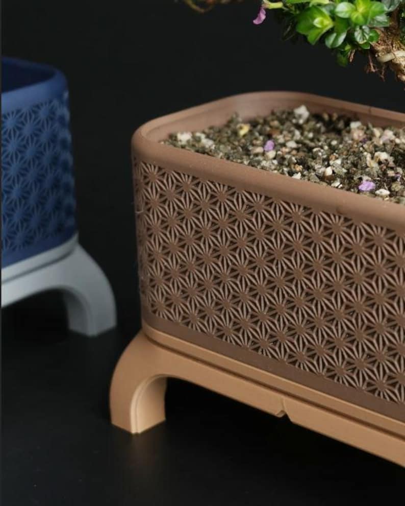 May include: A brown rectangular planter with a geometric starburst pattern, filled with soil and small stones. The planter sits on a matching base with curved legs. A blue planter with a similar design is visible in the background.
