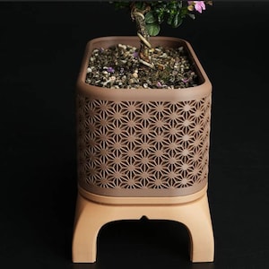 May include: A brown rectangular planter with a geometric star pattern, holding soil and a small plant with green leaves and pink flowers. The planter sits on a light brown base with curved legs.