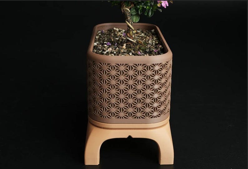 May include: A brown rectangular planter with a geometric star pattern, holding soil and a small plant with green leaves and pink flowers. The planter sits on a light brown base with curved legs.