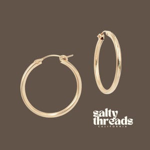 May include: Two gold-colored hoop earrings. The earrings are round and have a smooth, polished finish. The text "salty threads CALIFORNIA" is in white script on the bottom right corner of the image.