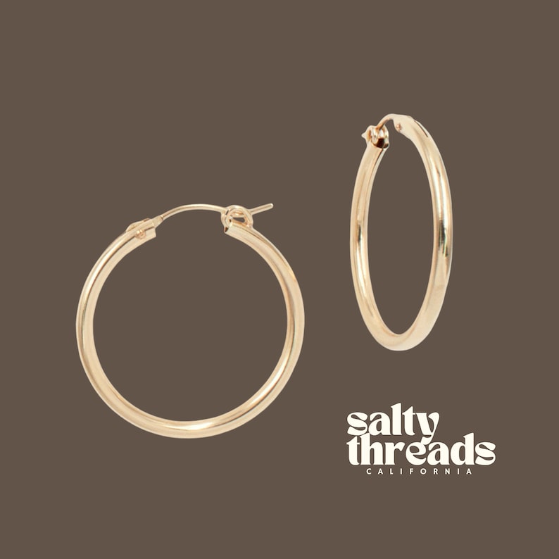 May include: Two gold-colored hoop earrings. The earrings are round and have a smooth, polished finish. The text "salty threads CALIFORNIA" is in white script on the bottom right corner of the image.