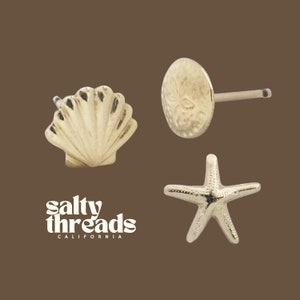 May include: Three gold-toned earrings featuring a seashell, a starfish, and a sand dollar. The earrings are small and delicate, perfect for adding a touch of beachy style to any outfit.