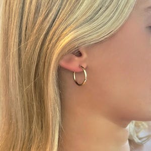 May include: A gold hoop earring is worn in the ear of a person with blonde hair. The earring is a simple, circular design, reflecting light. The earring is a classic jewelry piece.