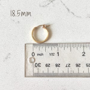 May include: A gold-colored hoop earring with a diameter of 18.5mm, next to a ruler showing inches. The earring is round and has a simple, classic design. The background is a light-colored, textured surface.