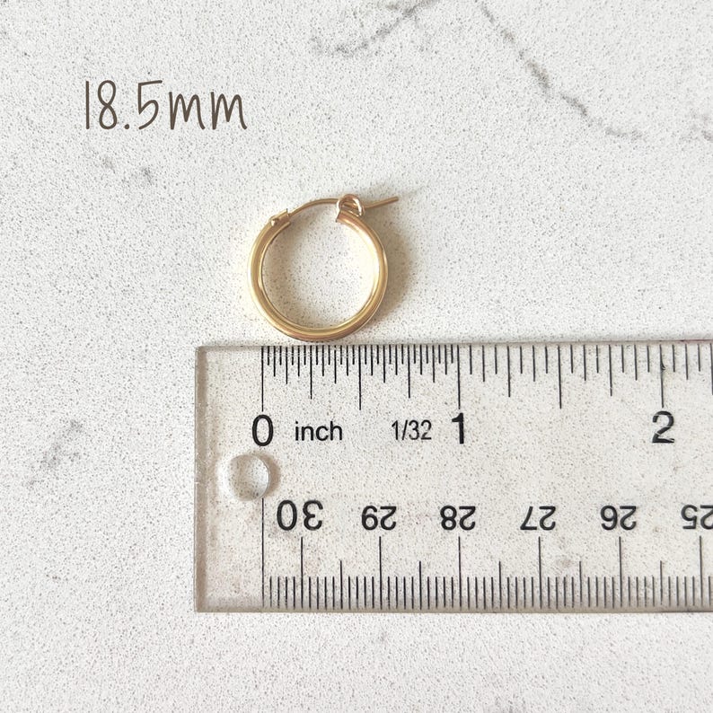 May include: A gold-colored hoop earring with a diameter of 18.5mm, next to a ruler showing inches. The earring is round and has a simple, classic design. The background is a light-colored, textured surface.