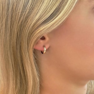 May include: A close-up of a gold hoop earring. The earring is small and round, with a polished finish. The earring is worn on the ear of a person with blonde hair.