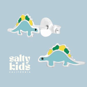 May include: A pair of silver stud earrings featuring a blue dinosaur with green and yellow accents. The earrings are from Salty Kids California.